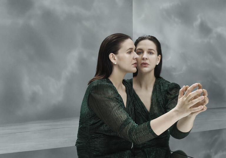 A woman on a grey stage leans against a mirror, her face reflects back at her. 