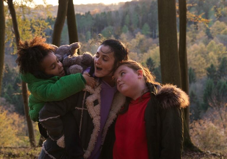 A mother holds her child in a green coat and hugs her elder daughter to her side, in the woods