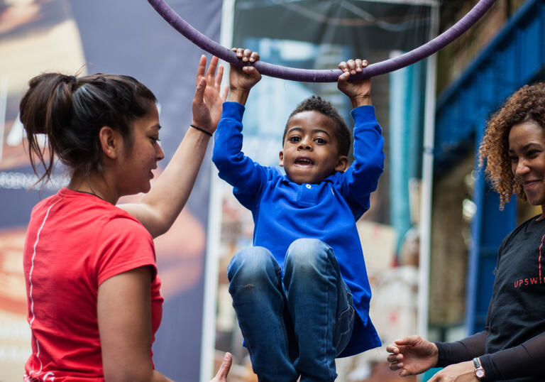 A child hanging from an aerial hoop with support from two workshop facilitators
