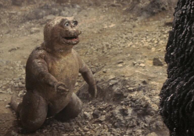 A film still showing a baby Godzille kneeling on the ground