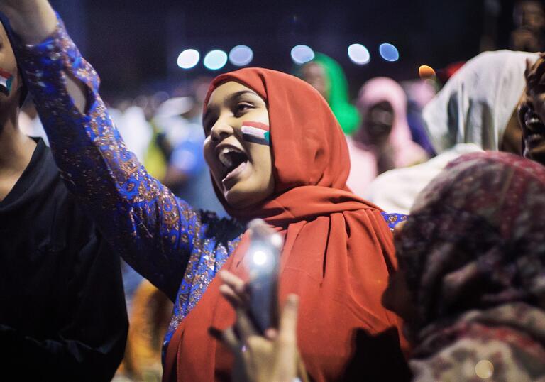 A woman in a red hijab and Sudanese flag on her face raises her hand in the air