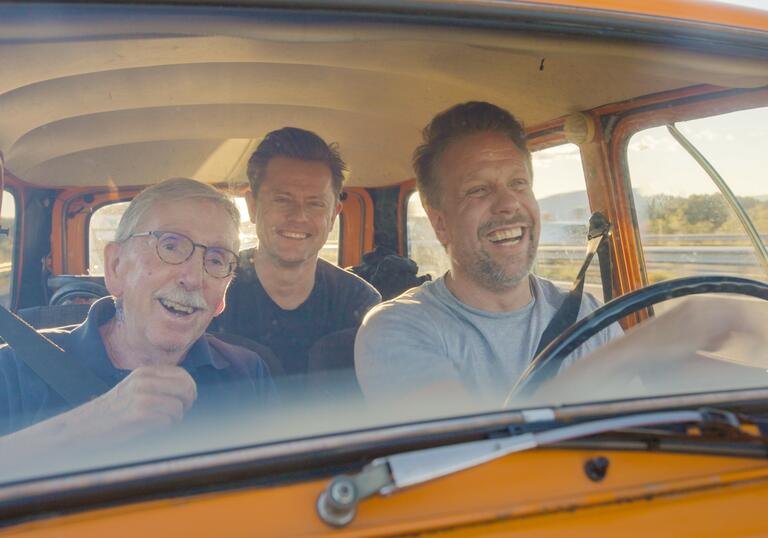 Three men sit in a yellow car, smiling