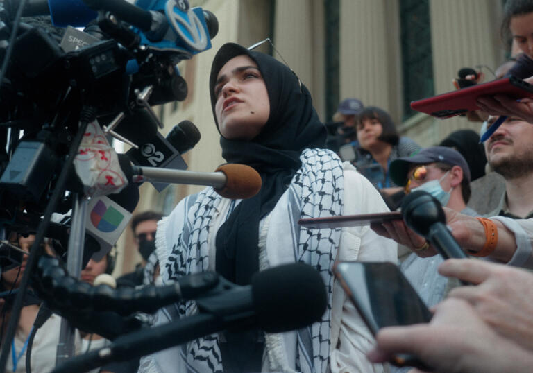 A woman wrapped in a kefiyah speaks into a camera and surrounded by journalists