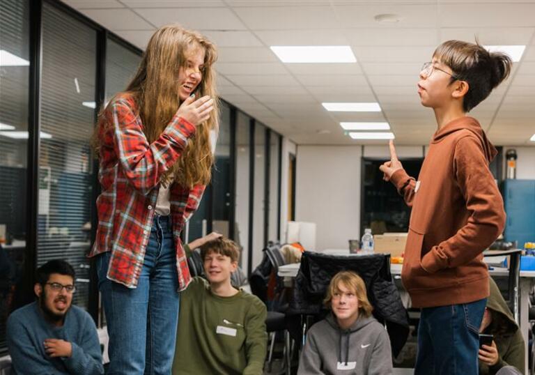 A group of children watch two others perform, one laughs as the other holds character.