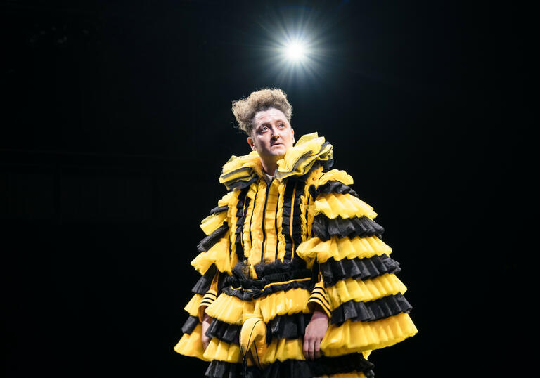 Malvolio stands on stage in a layered black and yellow costume, a light shines behind him.