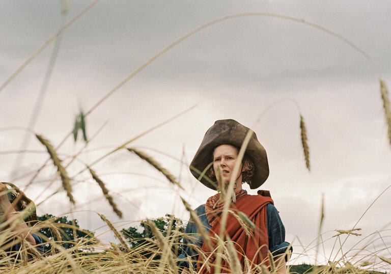 A person wearing a large leather hat and traditional Scottish tartan clothes stands obscured in a wheat field