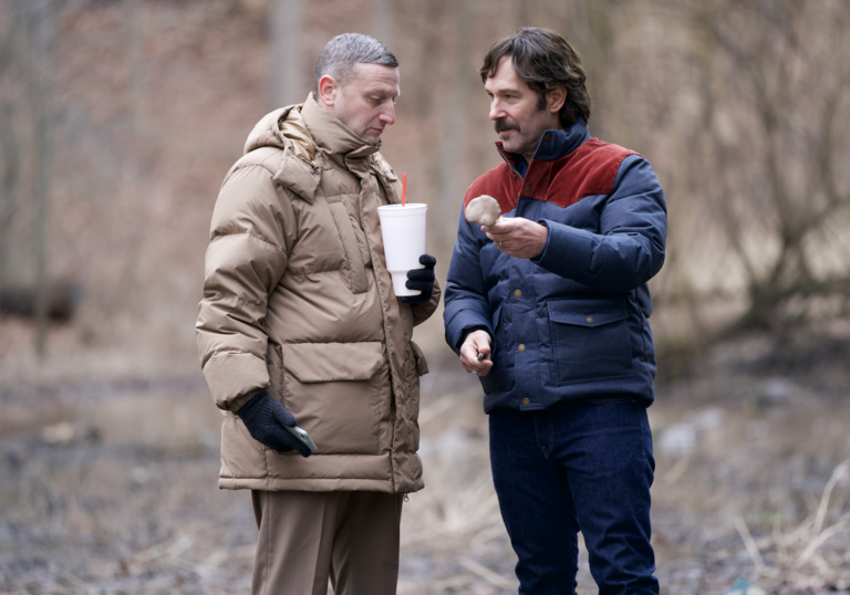 Two men in big winter coats share snacks on a walk