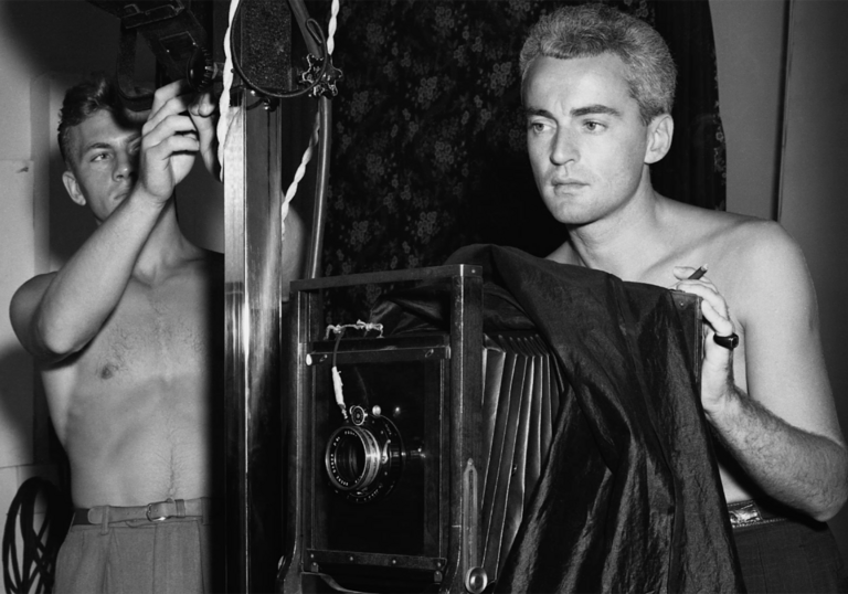 Two men stand behind an old fashioned camera, shirtless