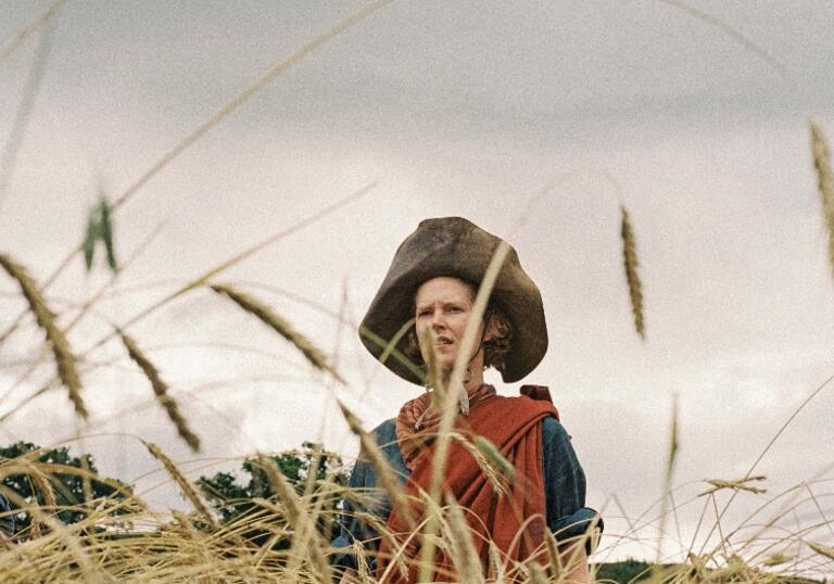 A woman wrapped in a red cloth and wearing a large leather hat stands in a field of wheat
