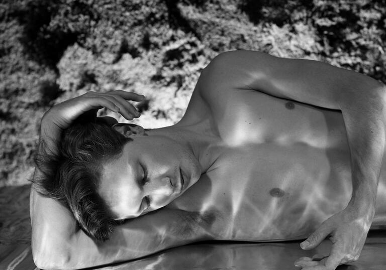 A topless white man lays on a mirror covered in water, staring at his reflection