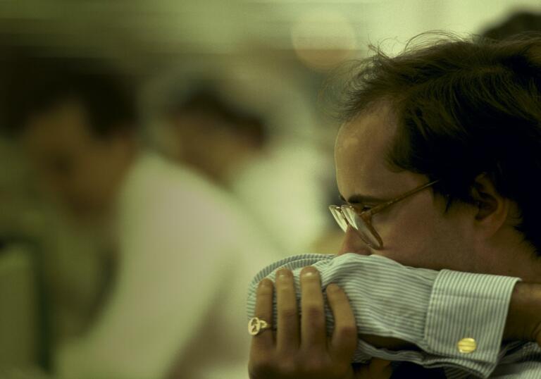 Man in smart clothing in glasses and with gold ring looks worried, covering his mouth with his arm