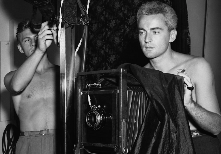 A man takes a photo of a man behind a camera, both are topless