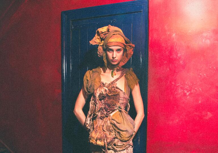 A model stands in front of a blue door and red wall, wearing earth toned distressed clothing