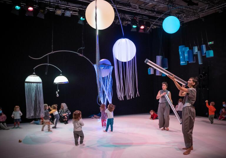 Toddlers running around with orbs of light hanging from the ceiling while two people overlook them