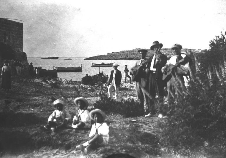 Black and white photo of Malta. There are three mail musicians performing and three children in the foreground. In the background is the ocean with some boats in it.