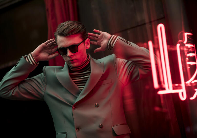 A man in glassed standing in front of a neon sign 