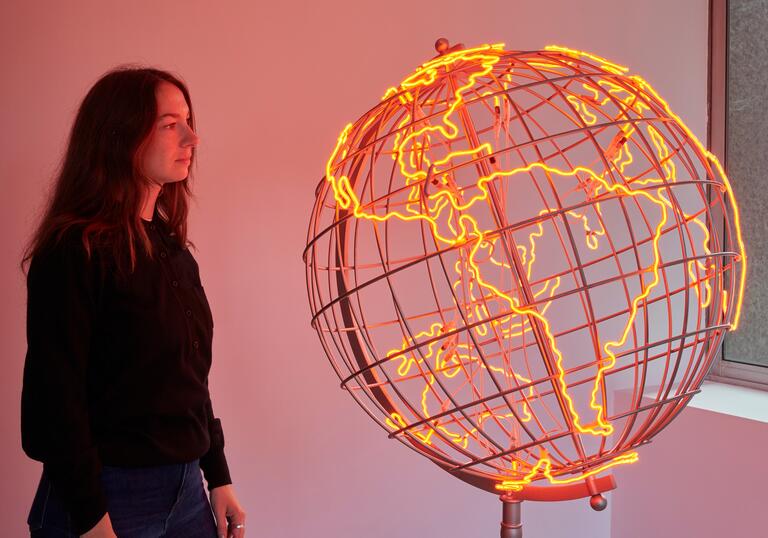 A woman looks at a sculpture of a globe, the outlines of the countries lit in neon orange strip lights.