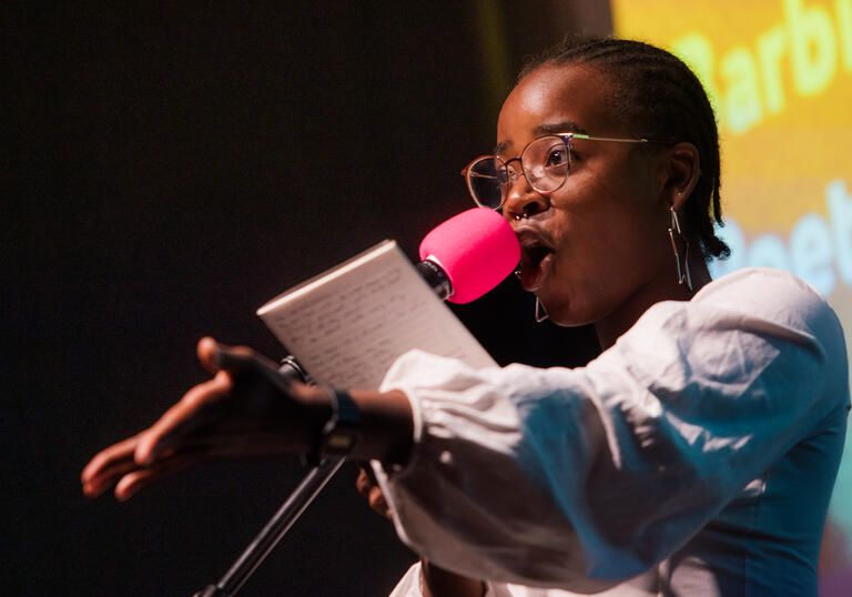 A young Black woman performing on stage speaking into a pink microphone, with her arm extended as she holds a book or paper.
