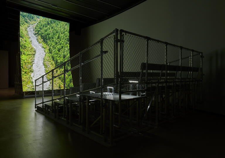 In a dark gallery, metal bleacher-style seating is positioned in front of a large, vertical, curved screen showing a river coursing through a lush, forested valley.