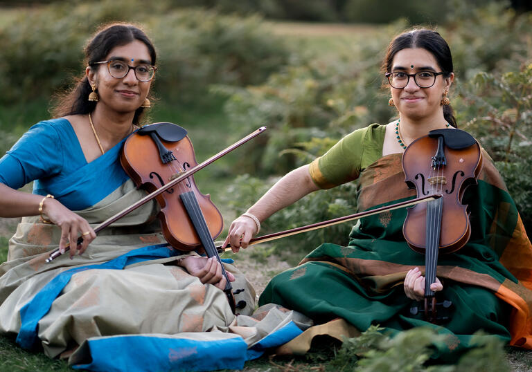 A photo of sibling duo Mathangi & Sambavi Uthayakumar sitting with violins on the grass.