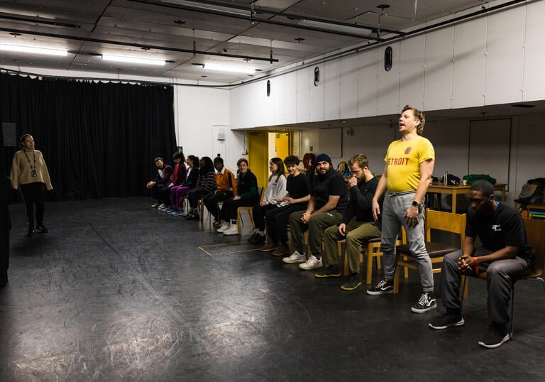 A group of workshop participants sit in a line of chairs. One of them is standing up and performing