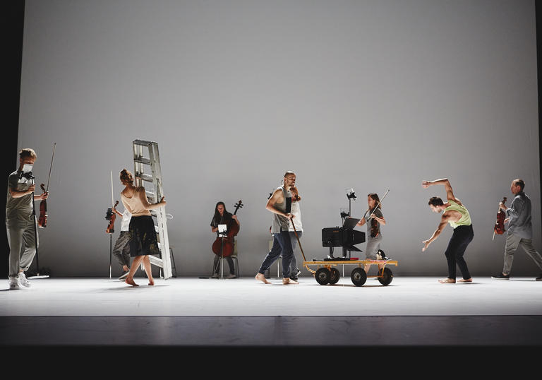 A photo of dancers and musicians in casual clothing on a stage with a white floor and a white background. Various musicians are holding different instruments including a cello and violins. A ladder is also being taken across the stage. A trolley with a stage light is being wheeled away.