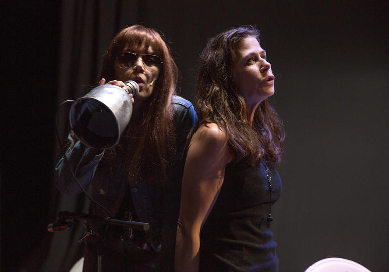 Image shows two women with long hair leaning against each other and singing