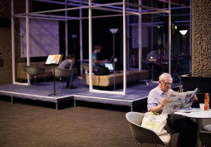 Photo of old man reading newspaper in the Barbican foyers during Hack the Barbican
