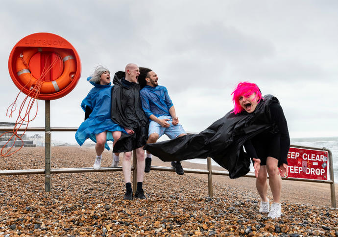 4 people in anoraks are on a windy beach. One has a bright pink wig. 