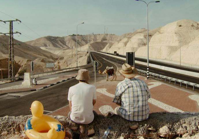 Two men sit on a wall looking out into the road into the distance, where a camel is standing