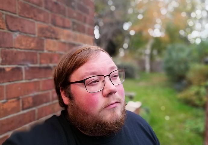image of artist Luke standing in front of a brick wall in a garden. Luke has a beard and glasses.