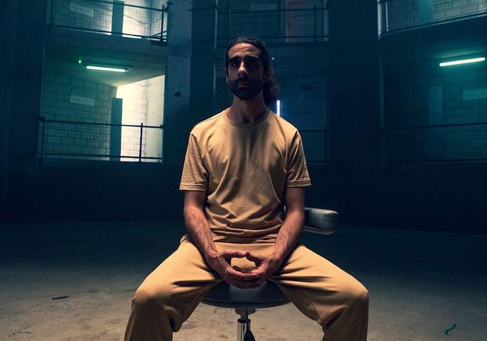 person sitting in dark room on a chair wearing beige t-shirt and trousers