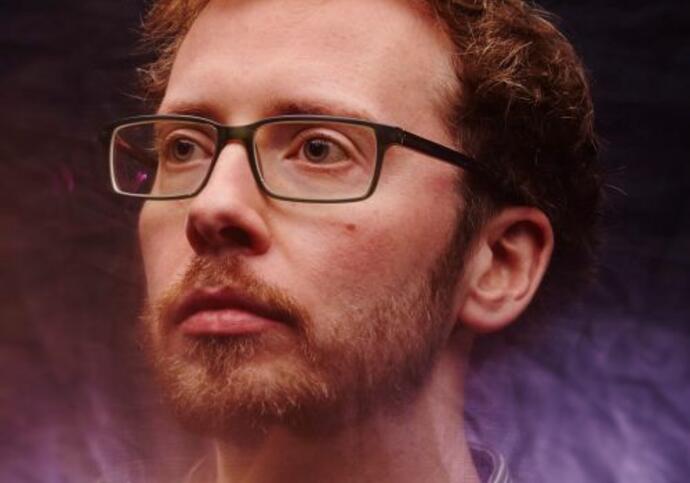Chris is standing in the rain with a spotlight behind him, looking up to the sky. He is wearing a blue shirt