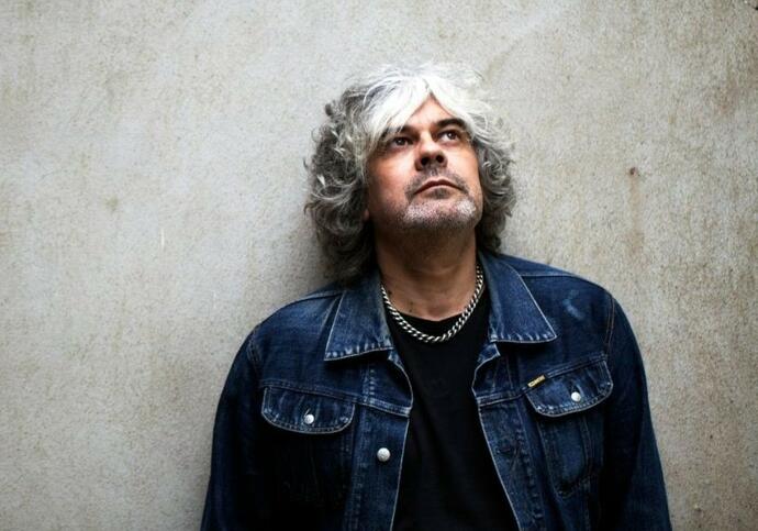 Matt is wearing a denim jacket and black t-shirt with a small chain around his neck. He is standing in front of a marble grey background.