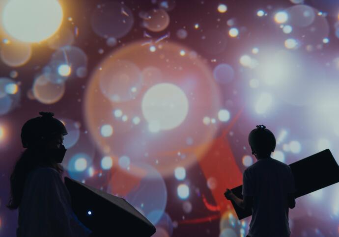 Two silhouettes of a girl and a boy wearing helmets and holding block shapes in front of a digital screen that shows colourful bubbles