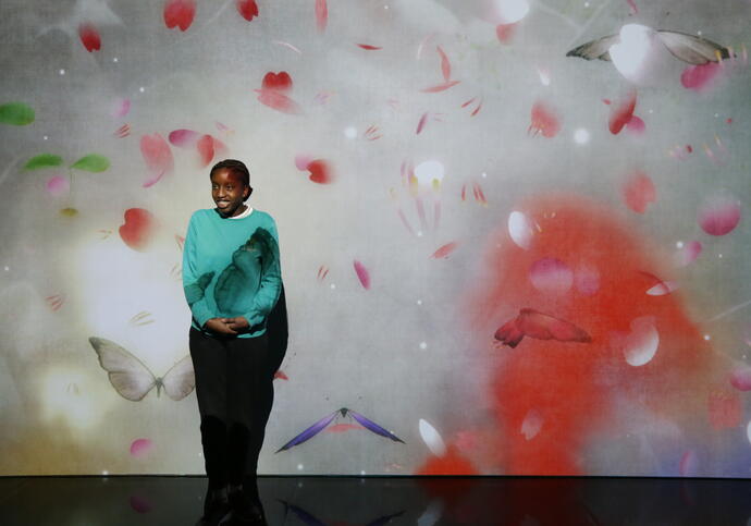 school child in uniform standing in front of a visual wall inside the exhibition. The wall has a projection of flowers floating in the air.