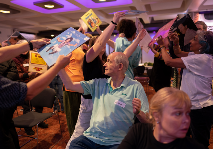 Group of people playing a game handing posters around, two people at the front sit while the rest behind them stand.