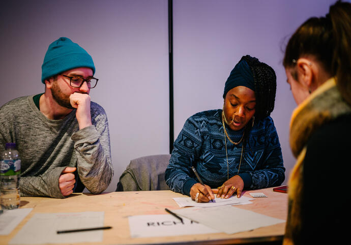 Three people sat at a table, one in grey jumper and blue beanie, another in blue jumper and another in black jumper and scarf