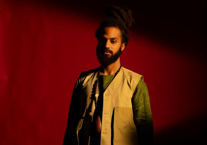 artist Edward is facing the camera standing against a dark red background. They are wearing a khaki t-shirt and beige vest. 
