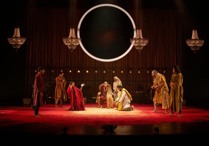 An ensemble of performers gather on stage in a decadent room, a large circle light hangs above them.