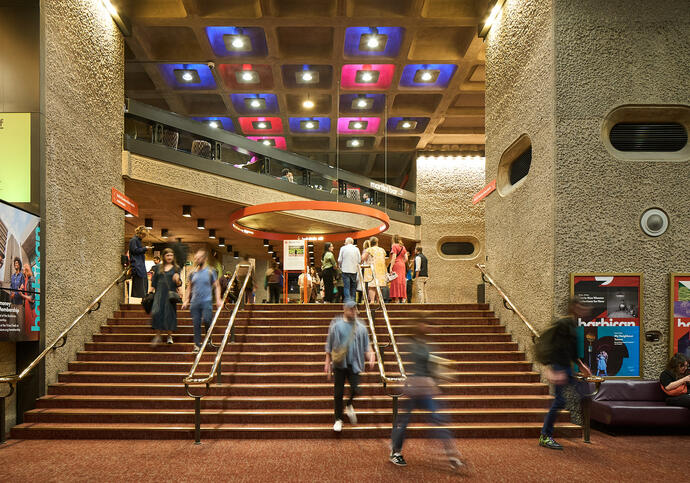 Blurred people walk down the stairs from the Barbican foyers, with bright ceiling panel lights