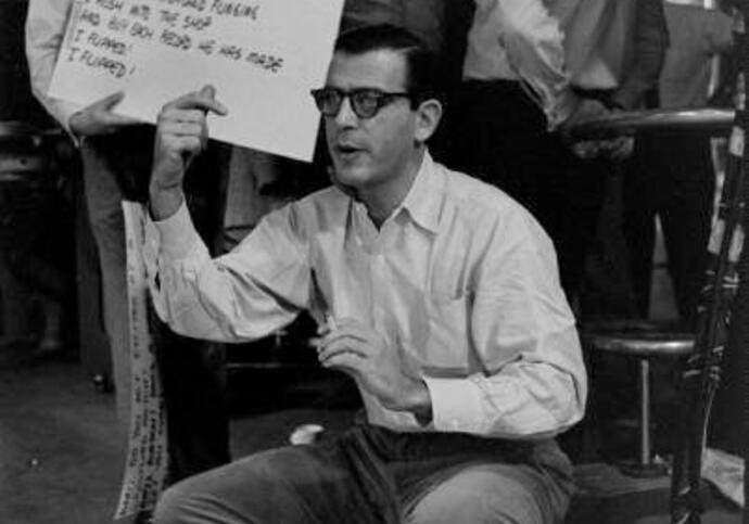 Black and white image of Oscar Levant squatting in front of a person with a sign
