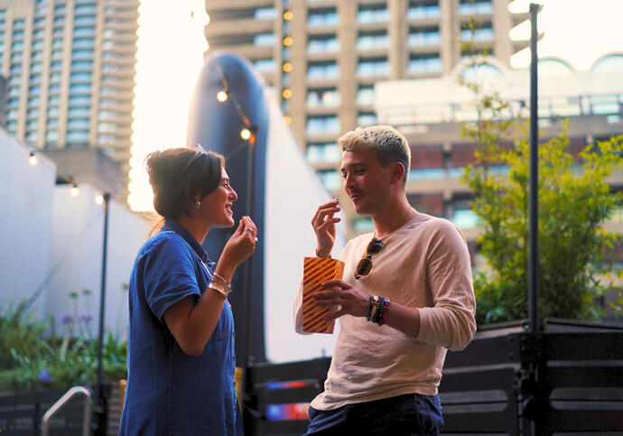 A man and a woman enjoy some popcorn outdoors. The sun is low in the sky approaching evening and they are surrounded by concrete buildings and some green foliage.