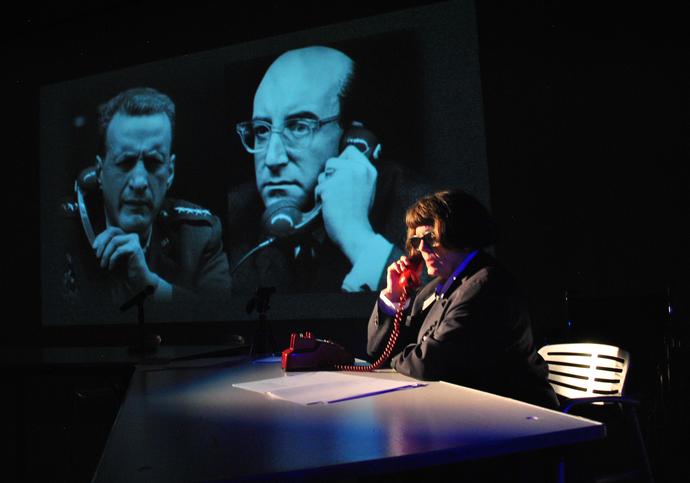 A photo showing a character called Lois Weaver in glasses, wearing a black jacket on the phone. It is a red coloured phone and is on a table with some paper. In the background there is a clip from Dr. Strangelove the film