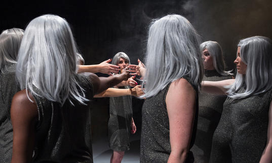 Photos which shows a group of women with identical silver wigs holding their hands out into a circle