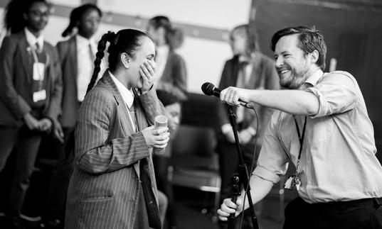 Photo of girl in school uniform laughing as teacher holds microphone