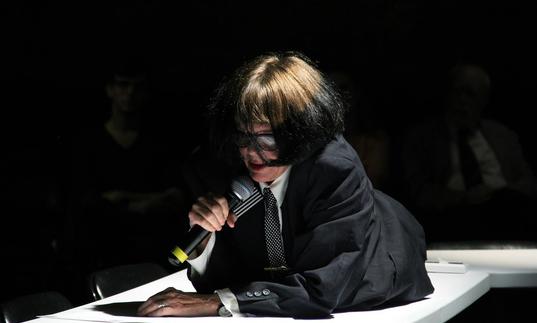 Photo of a woman in glasses using a mic. She is lying on a table. 