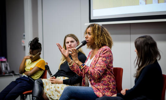 Four people on a panel, one talking into the microphone and gesticulating with a screen behind them.