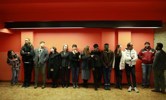 13 people standing in a line along an orange wall, talking and laughing