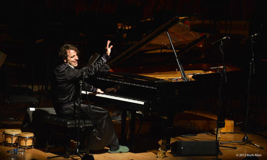 Chilly Gonzales is playing a piano one handed whilst doing a wave